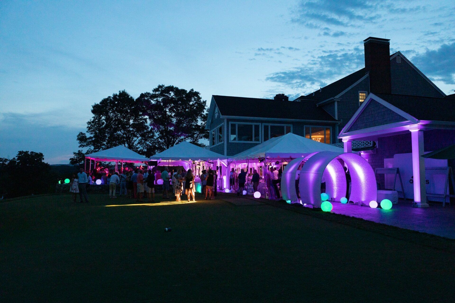 Outdoor evening event with purple-lit tents beside a large house, with guests gathered on the lawn