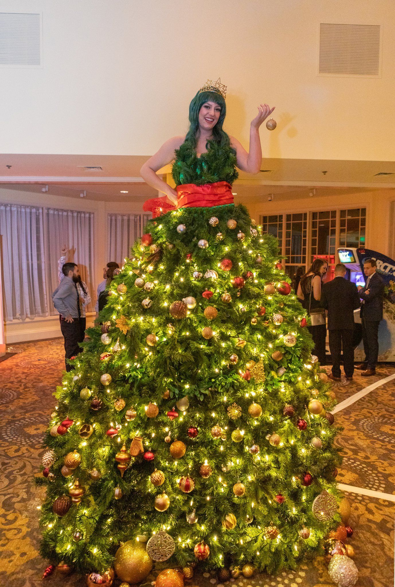 Person standing on a decorated Christmas tree costume in a hotel lobby with lights and ornaments