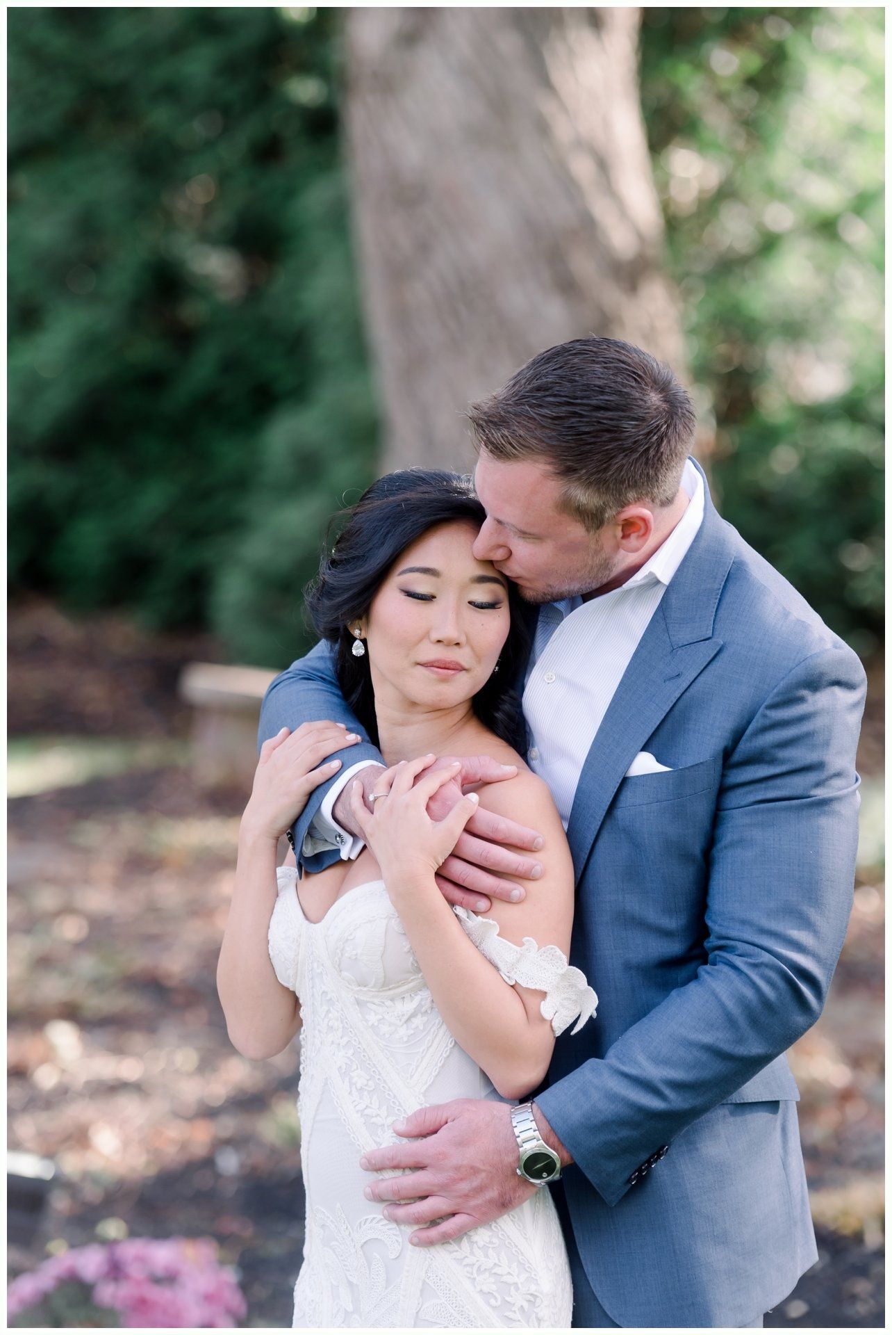 Bride and groom embracing outdoors beneath a tree, with the groom kissing her forehead.