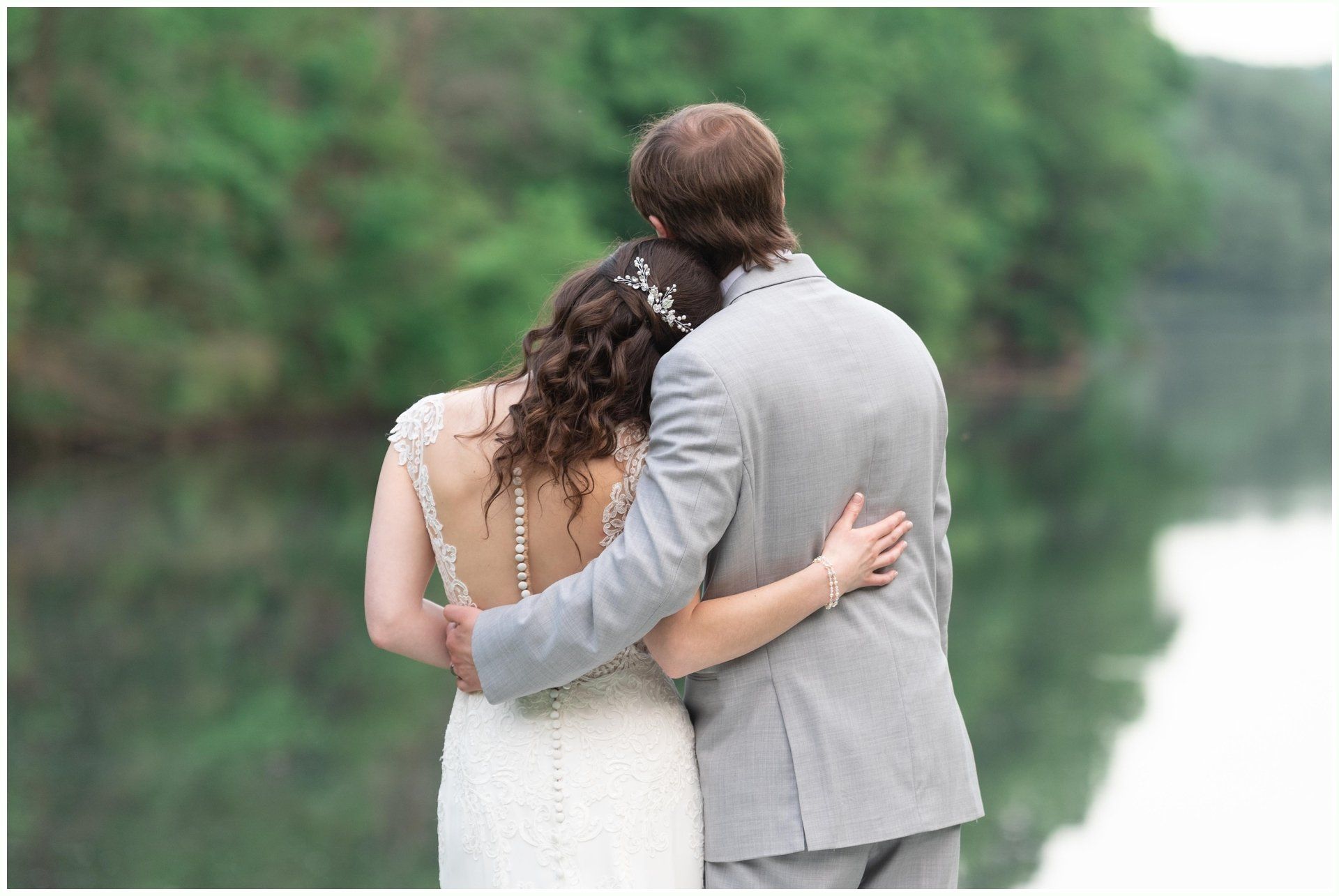 Couple in wedding attire embracing by a lake with green trees in the background