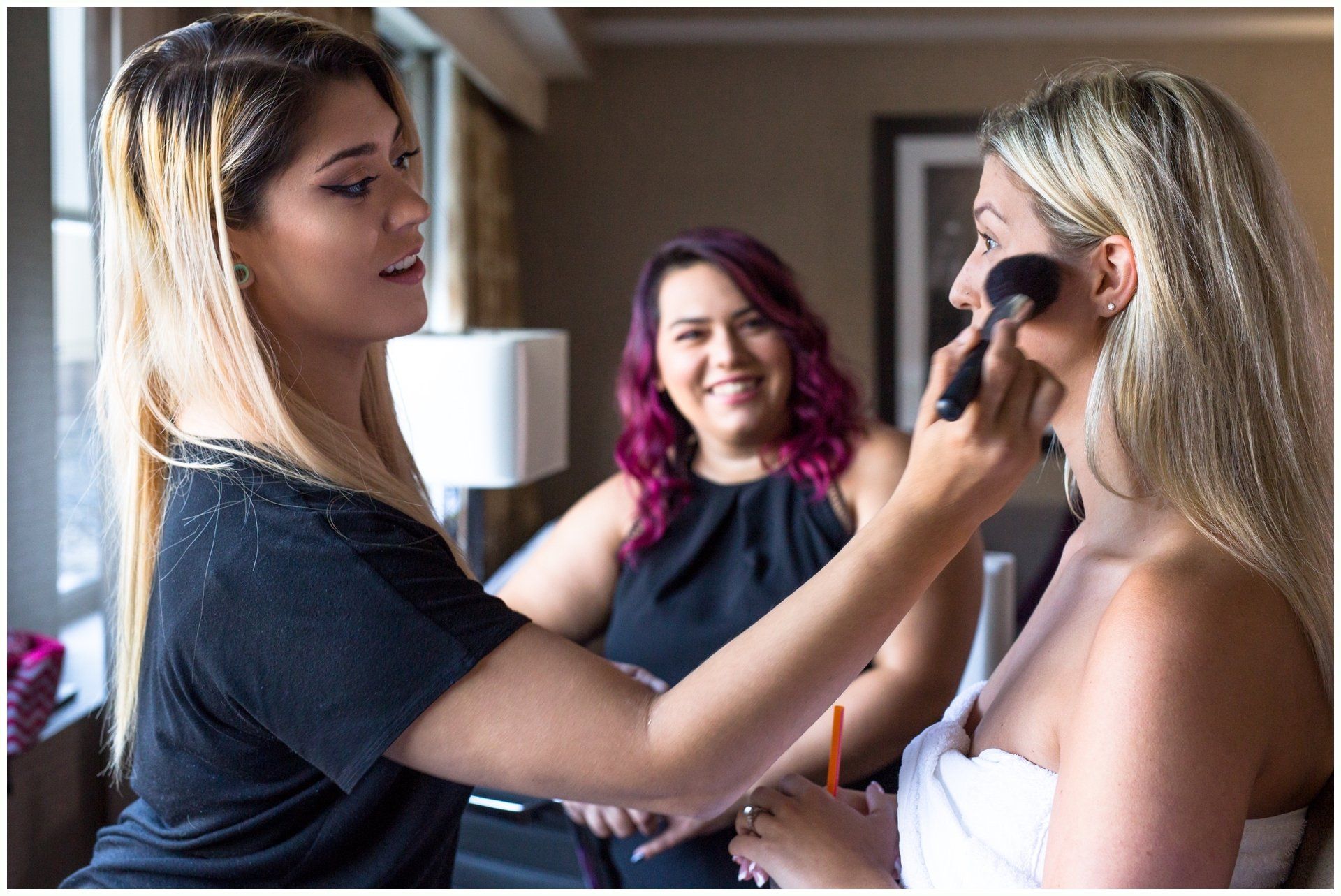 Makeup artist applying makeup to a smiling woman while another woman watches in a room
