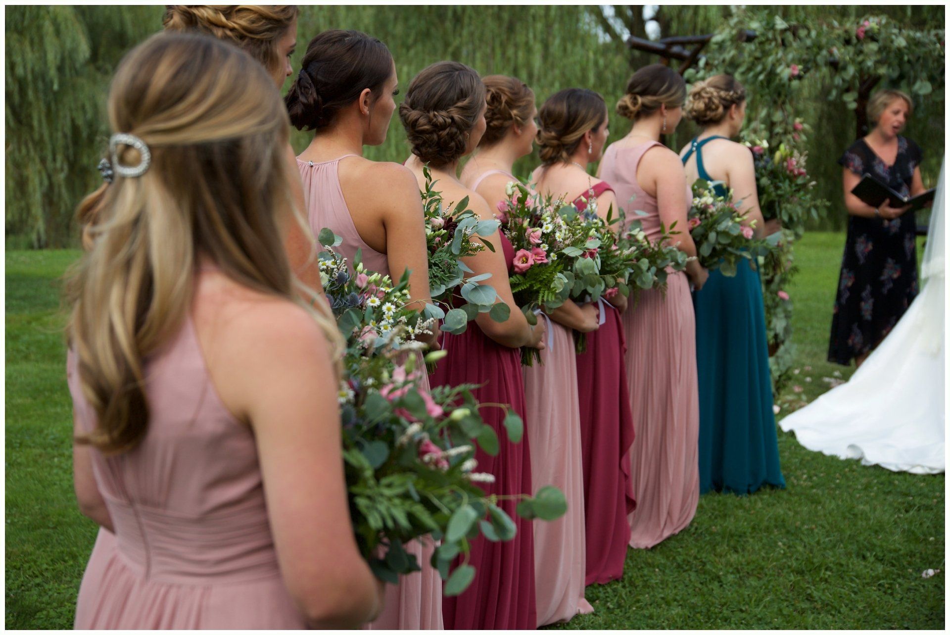 Bridesmaids in pink and teal dresses hold bouquets at an outdoor wedding ceremony.