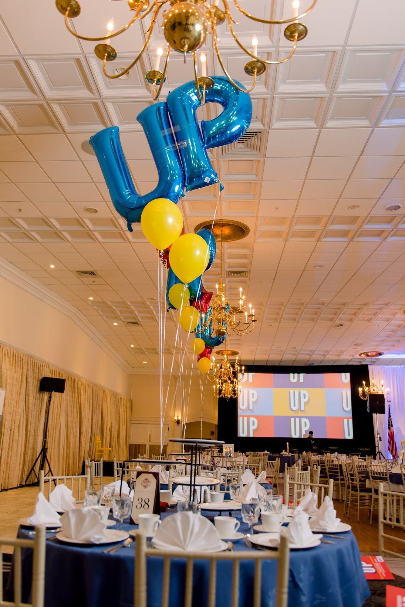 Banquet hall with blue and yellow balloon column, decorated tables, and a stage with colorful backdrop