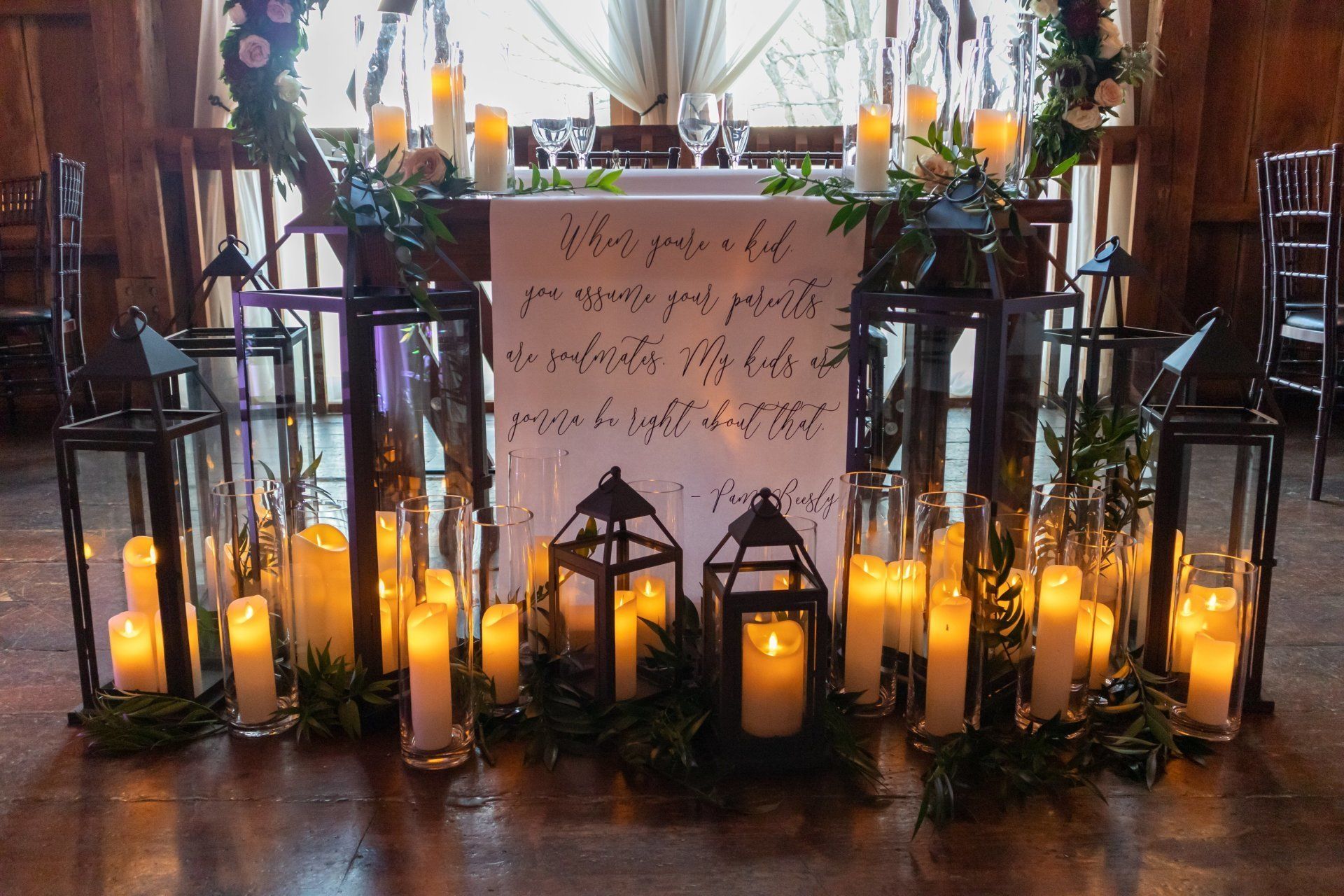 Candlelit wedding welcome display with lanterns, greenery, and a handwritten sign on a table