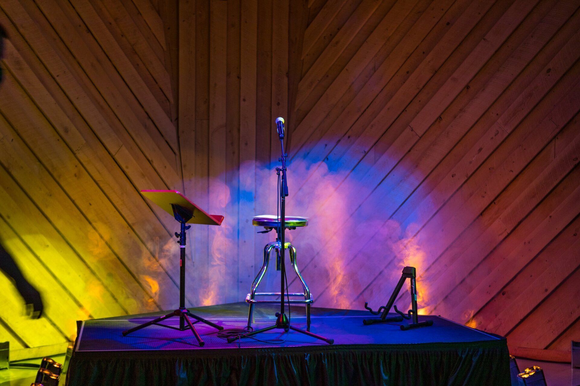 Empty drum kit on a small stage with colorful yellow and blue lighting and smoke against a wood-paneled wall