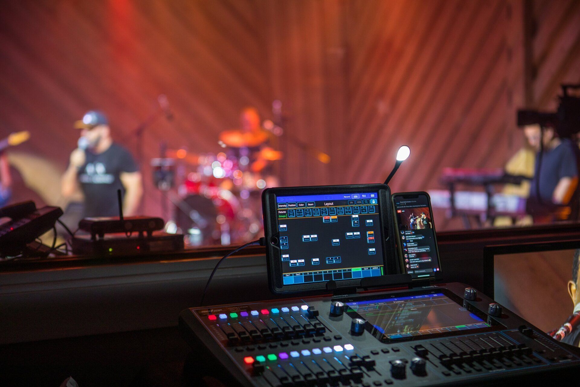 Sound mixing console with screens in foreground, band performing on a lit stage in the background.