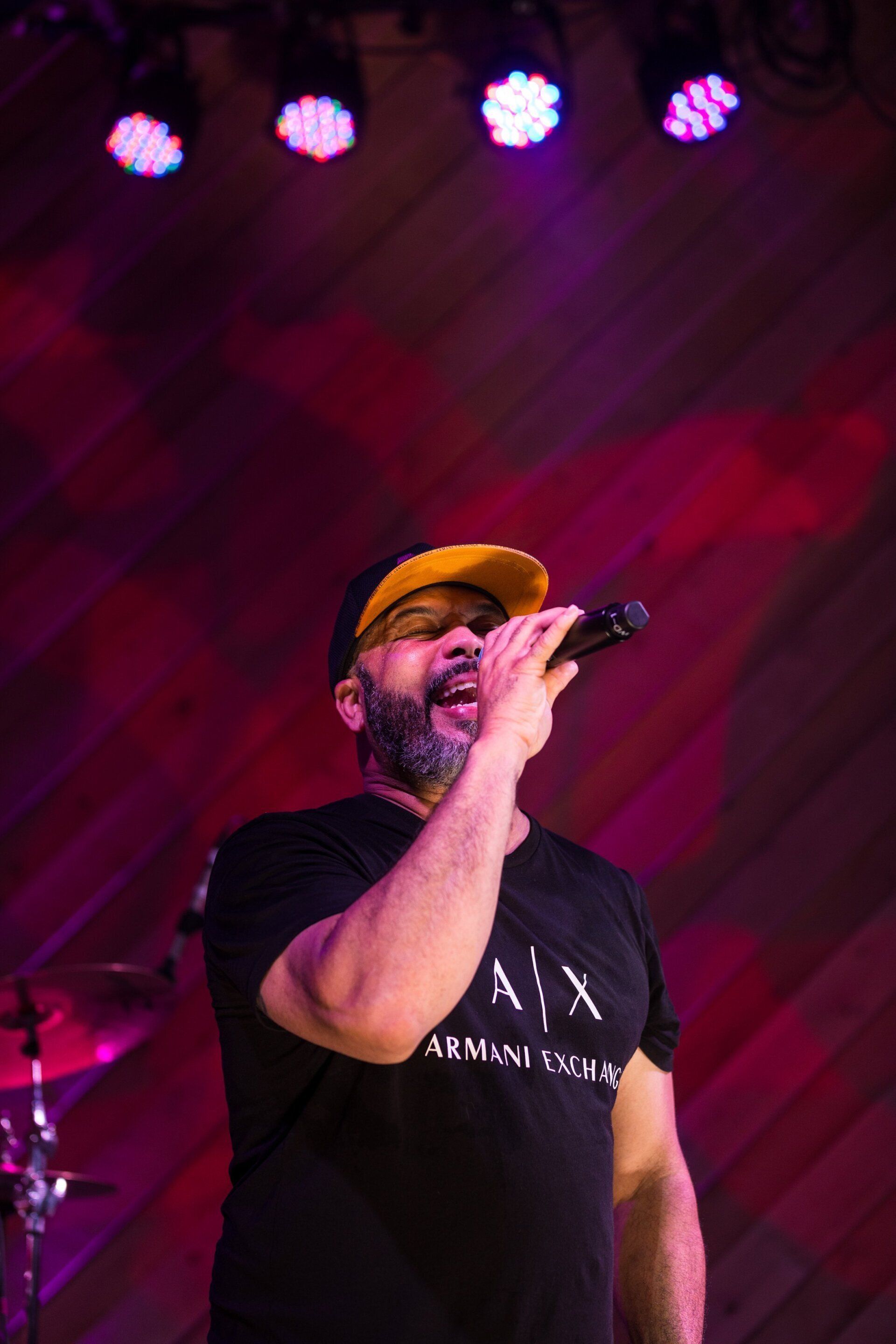 Singer in a yellow cap and black shirt performing onstage under pink and purple lights