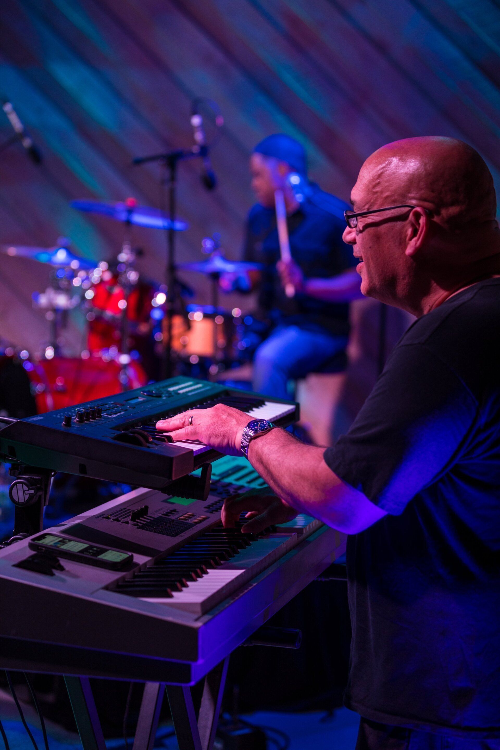 Musician playing keyboard on stage under blue and purple lights, with a drummer blurred in the background