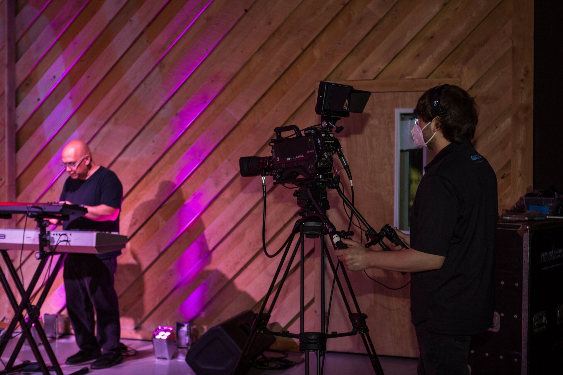 Camera operator filming a bald keyboard player onstage under purple lighting