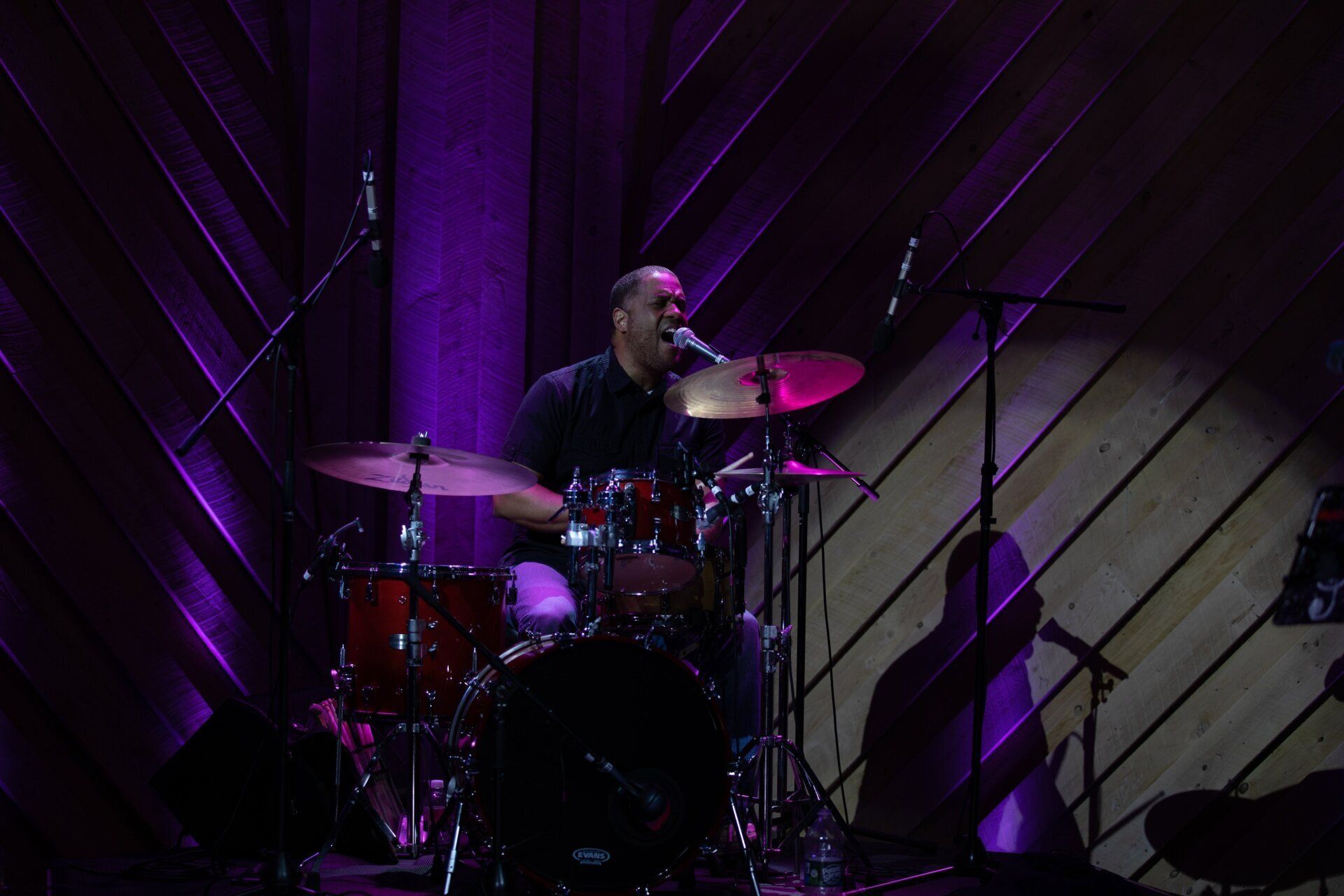 Drummer performing on a purple-lit stage with diagonal wooden wall panels behind him