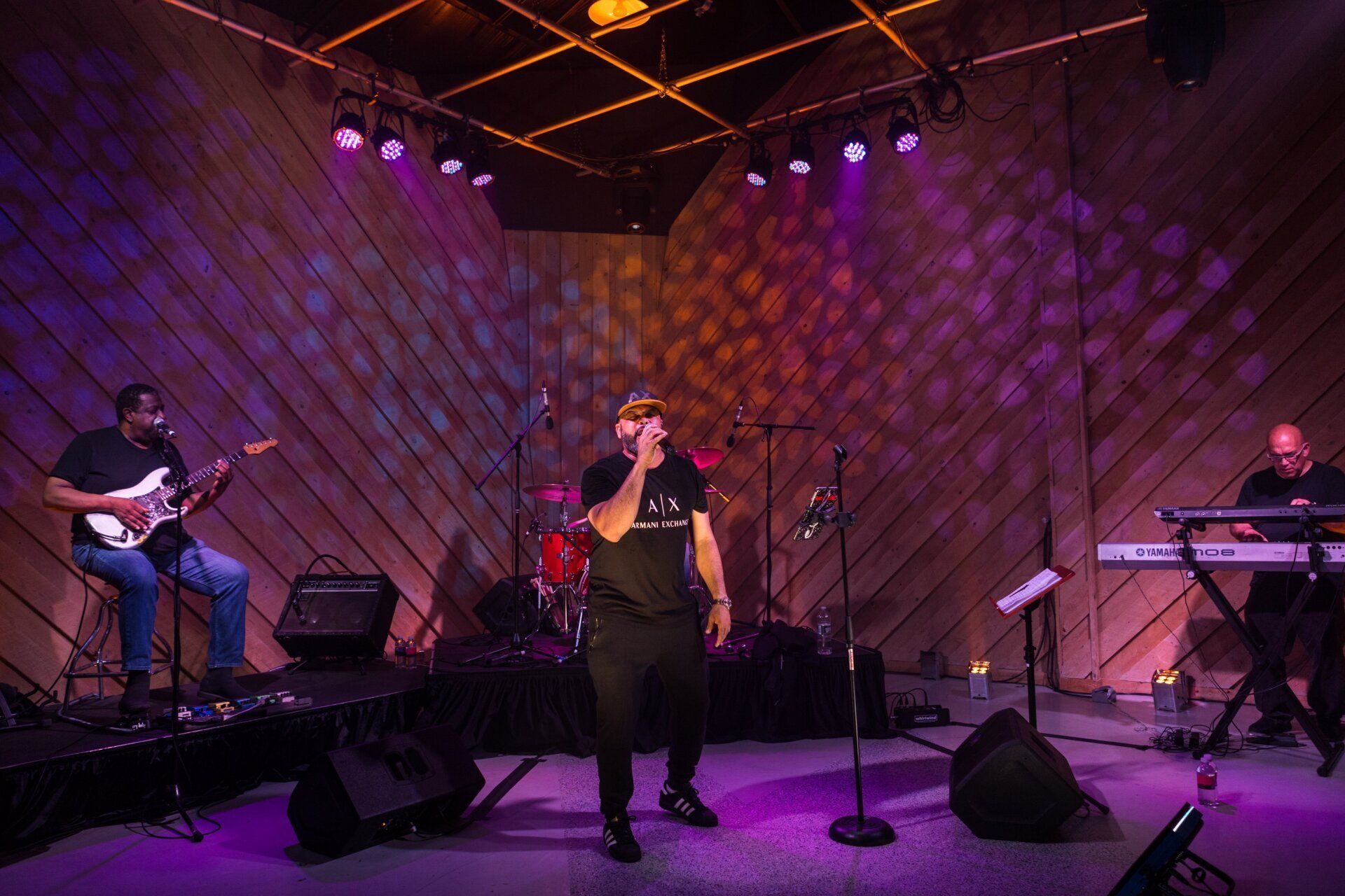 Live band performing on stage under purple lights, with singer center, guitarist left, and keyboardist right.