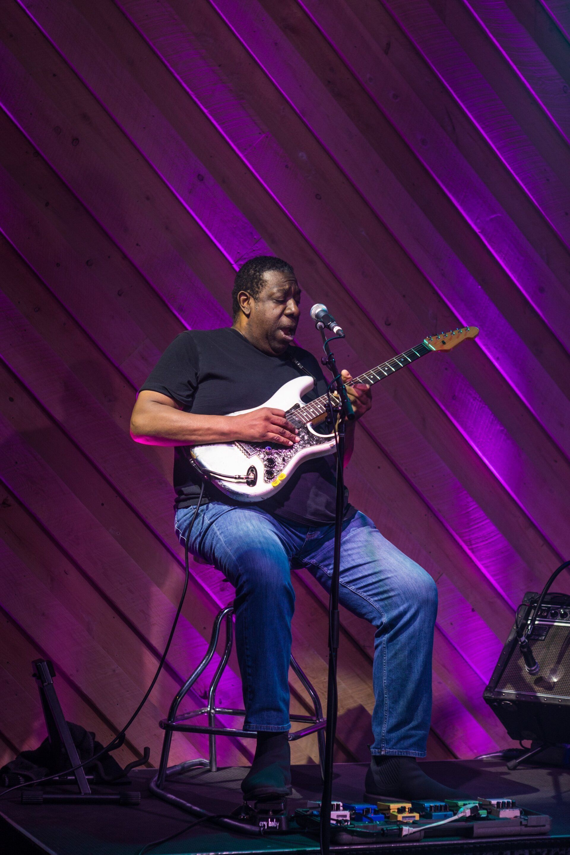 Musician seated on stage playing electric guitar under purple lighting