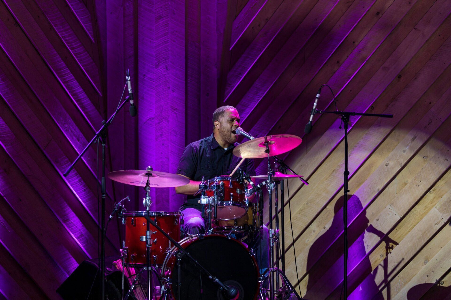 Drummer on stage under purple lighting, performing in front of a wooden backdrop.