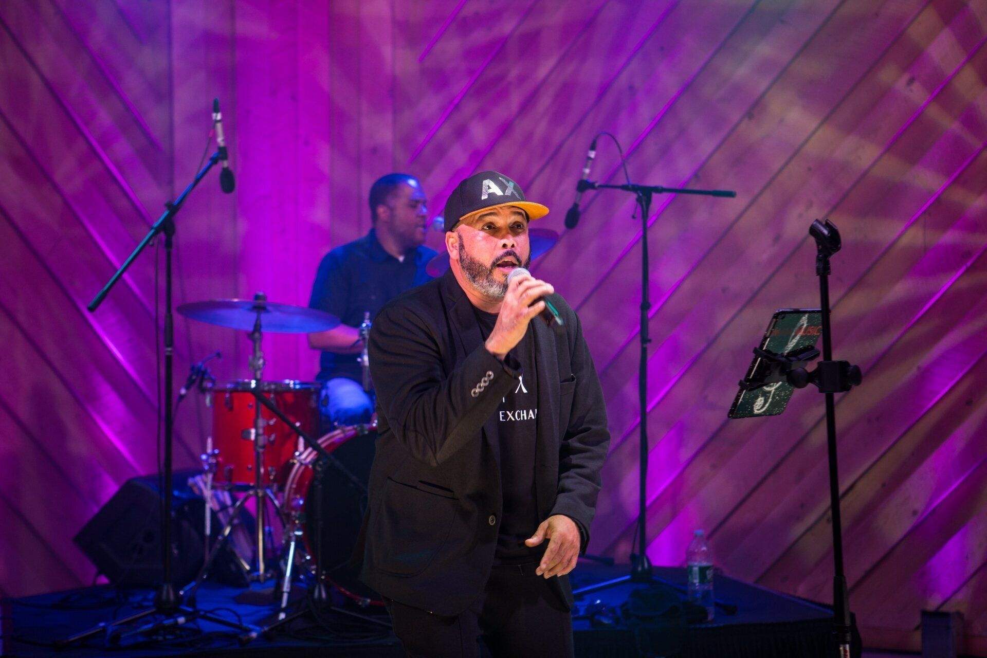 Singer performing on stage with drummer, purple-lit backdrop, holding a microphone