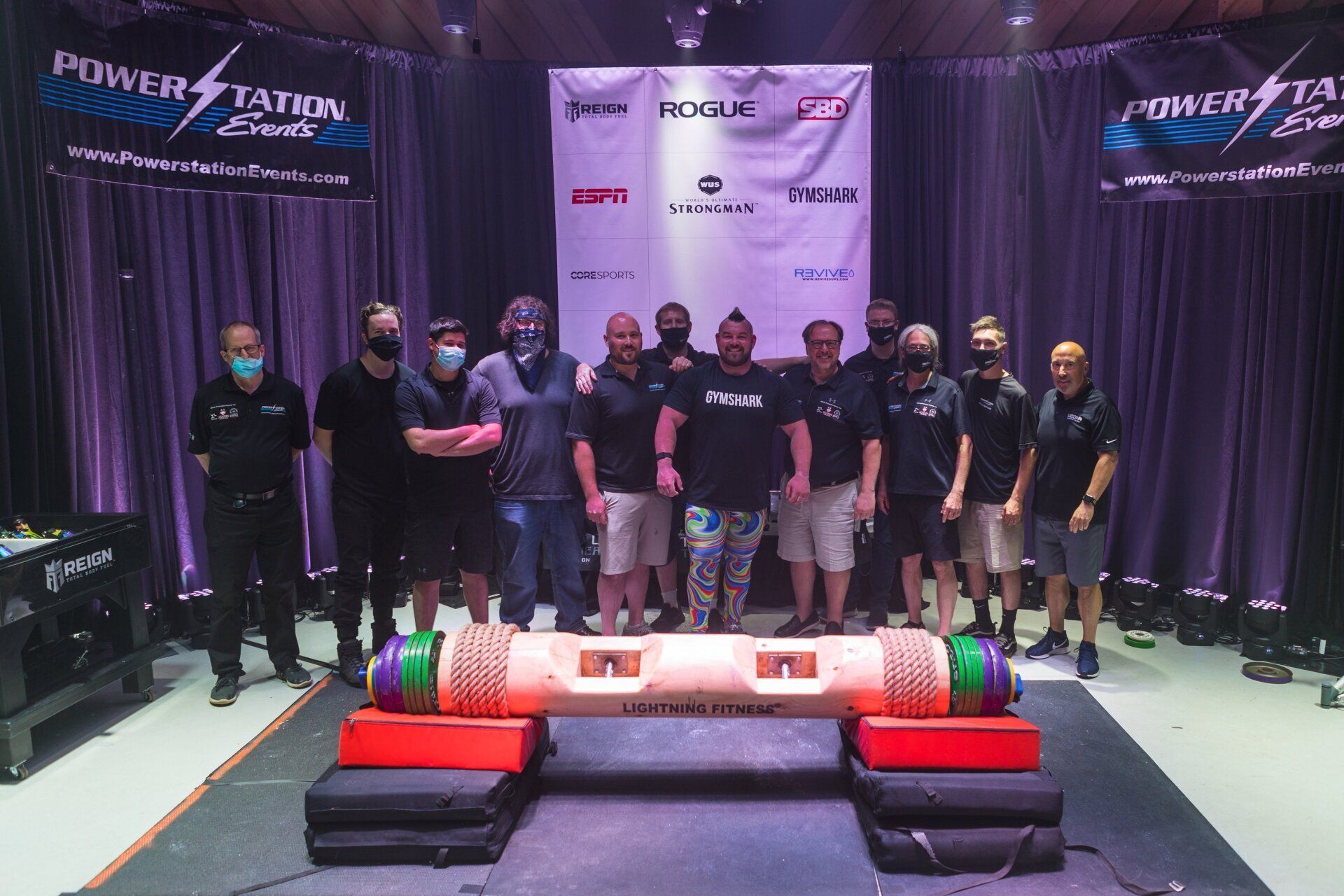 Group of people posing around a giant game controller on a stage with purple curtains and banners