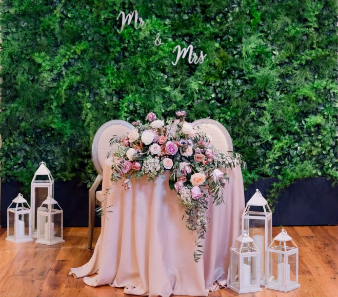 Pink floral wedding display with draped table, white lanterns, and “Mr & Mrs” on a green backdrop