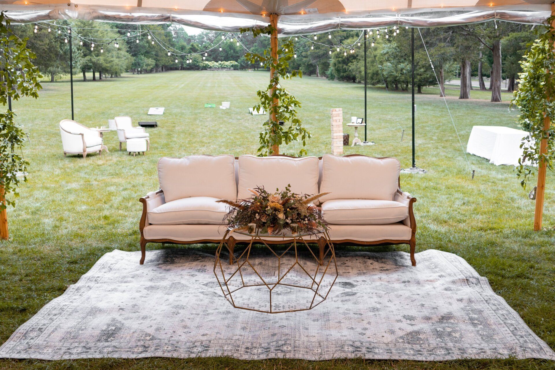 Outdoor lounge under a tent with a beige sofa, gray rug, and grassy yard beyond the clear walls