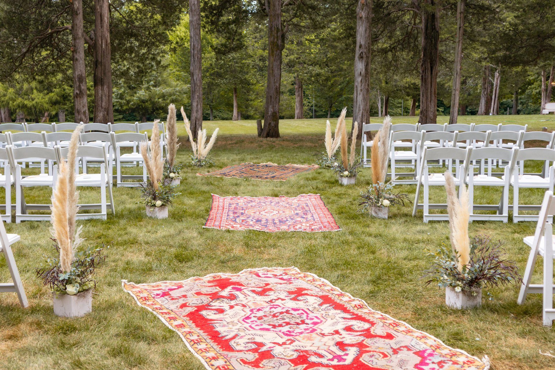Outdoor wedding aisle with white chairs, red patterned runners, and white floral arrangements in a grassy park