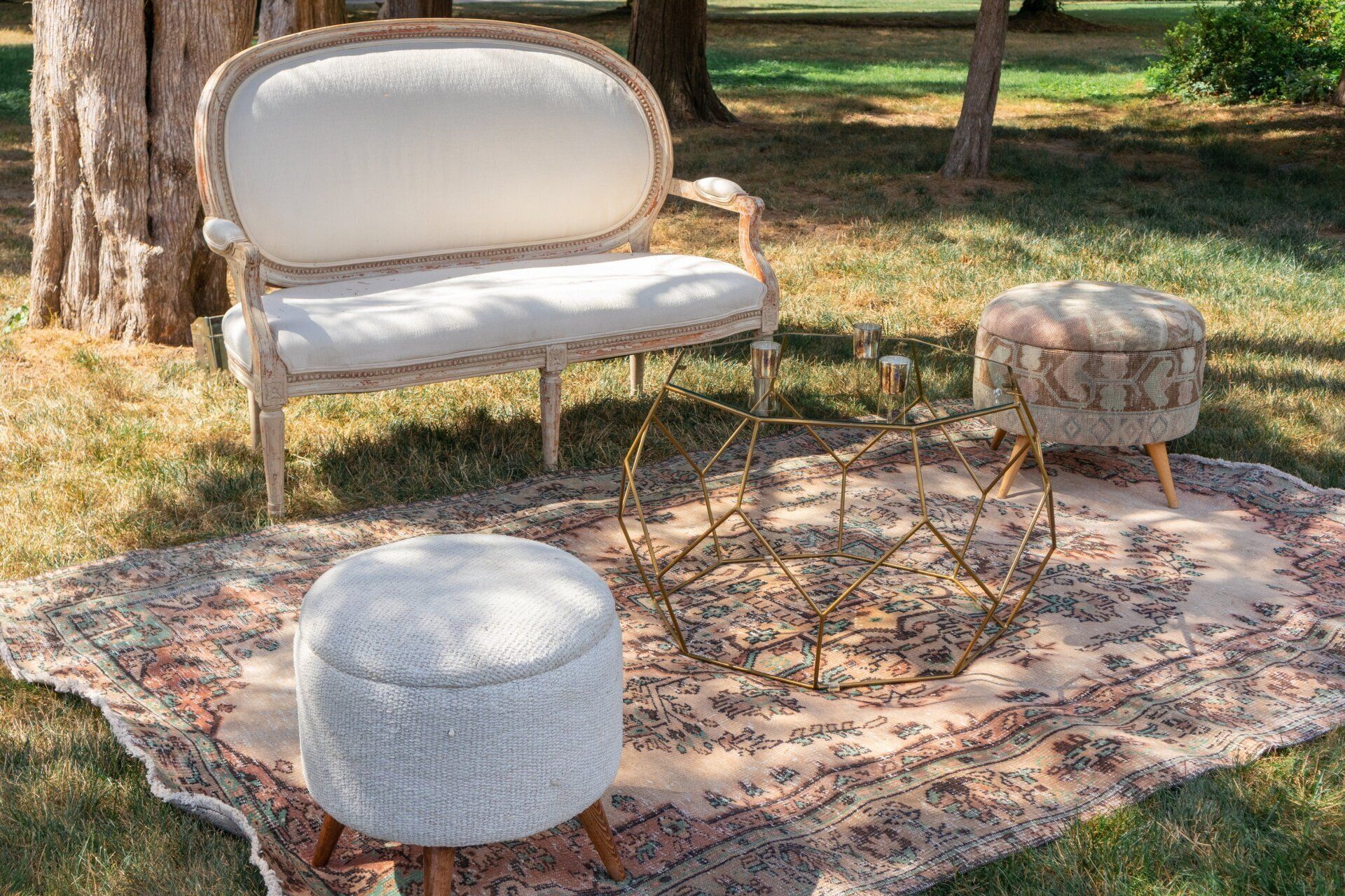 Vintage white loveseat and pouf on a patterned rug outdoors among trees and grass.