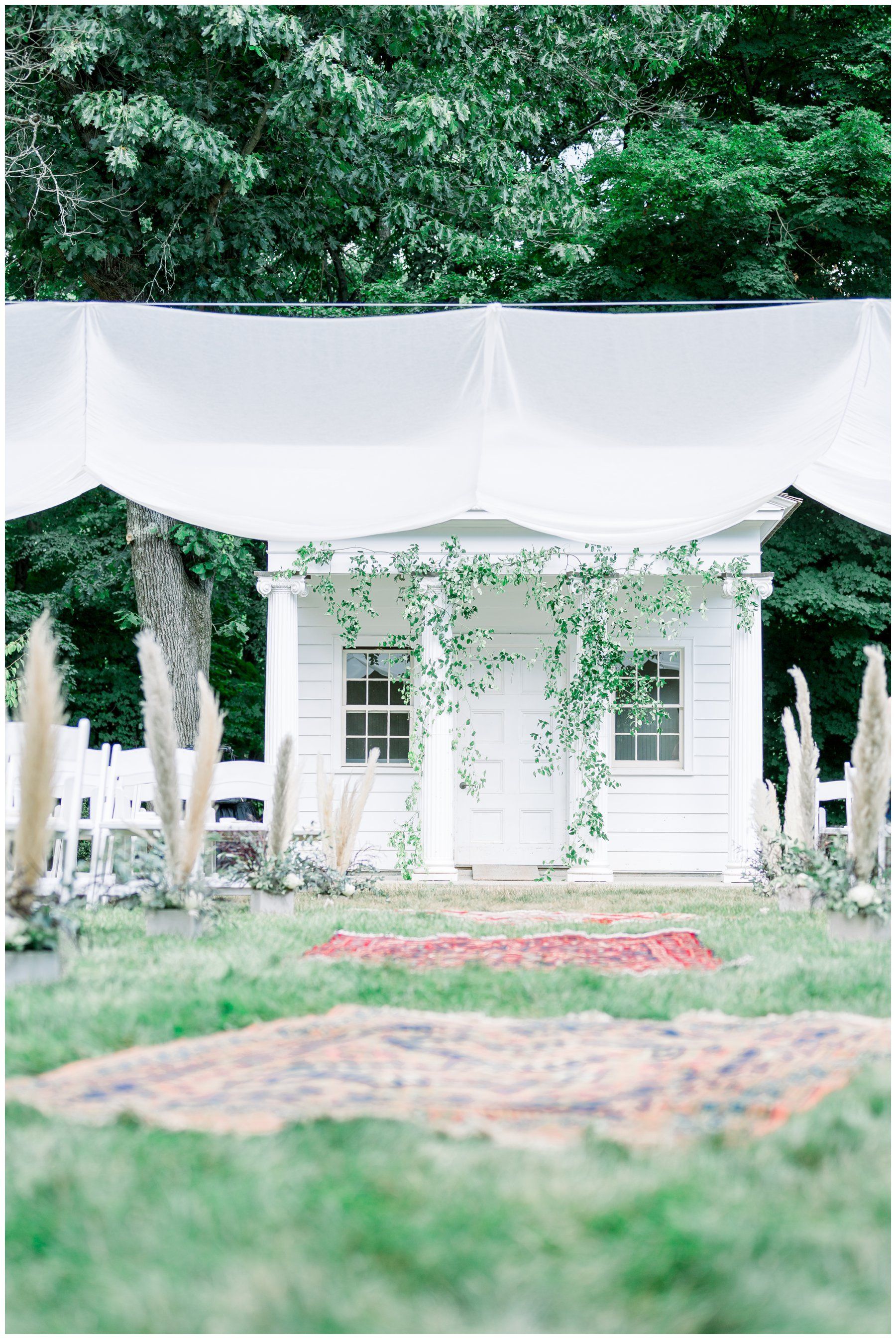 White draped outdoor wedding canopy in a lush green garden with chairs and a small white structure behind it