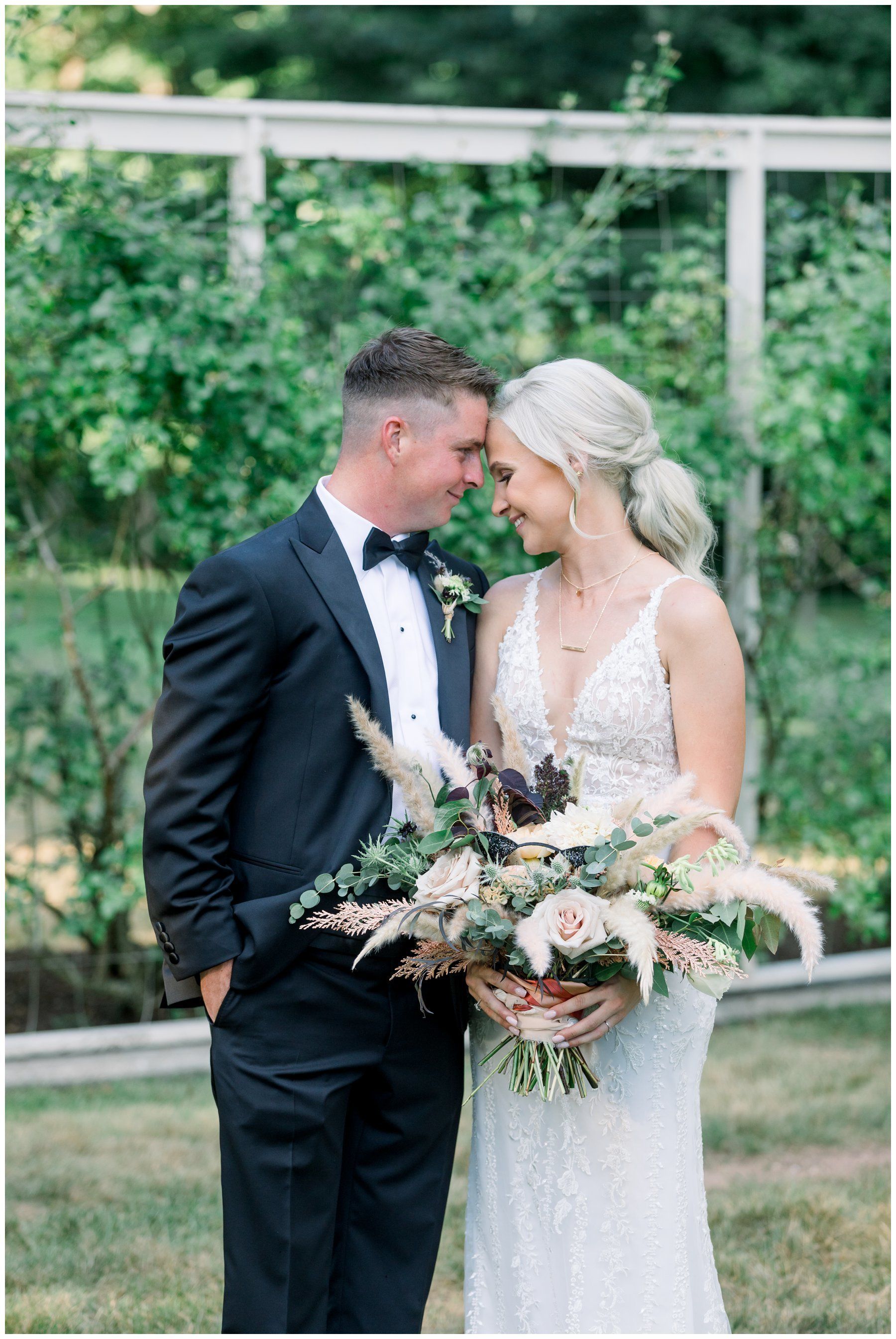 Bride and groom in formal attire sharing a forehead touch while holding a large floral bouquet outdoors