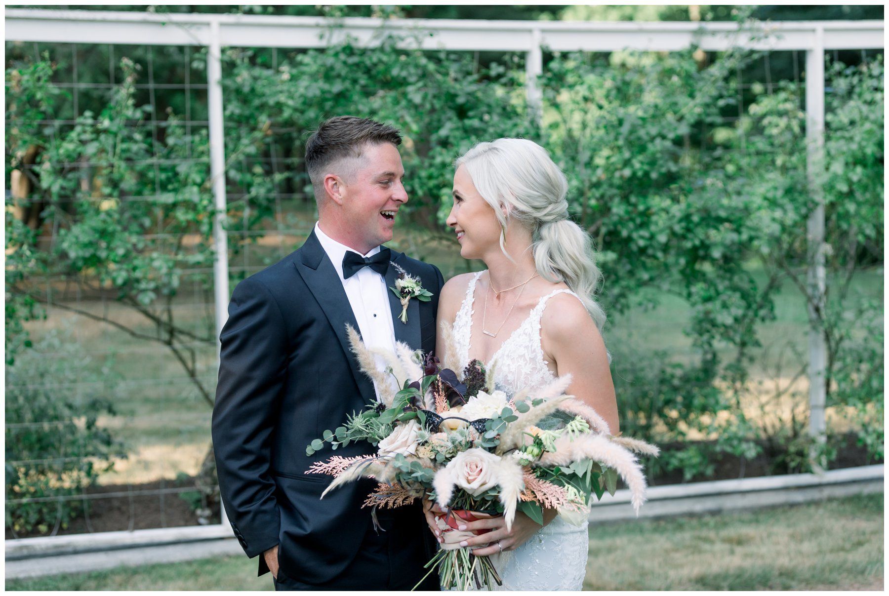 Couple in wedding attire smiling at each other outdoors, holding a bouquet amid greenery