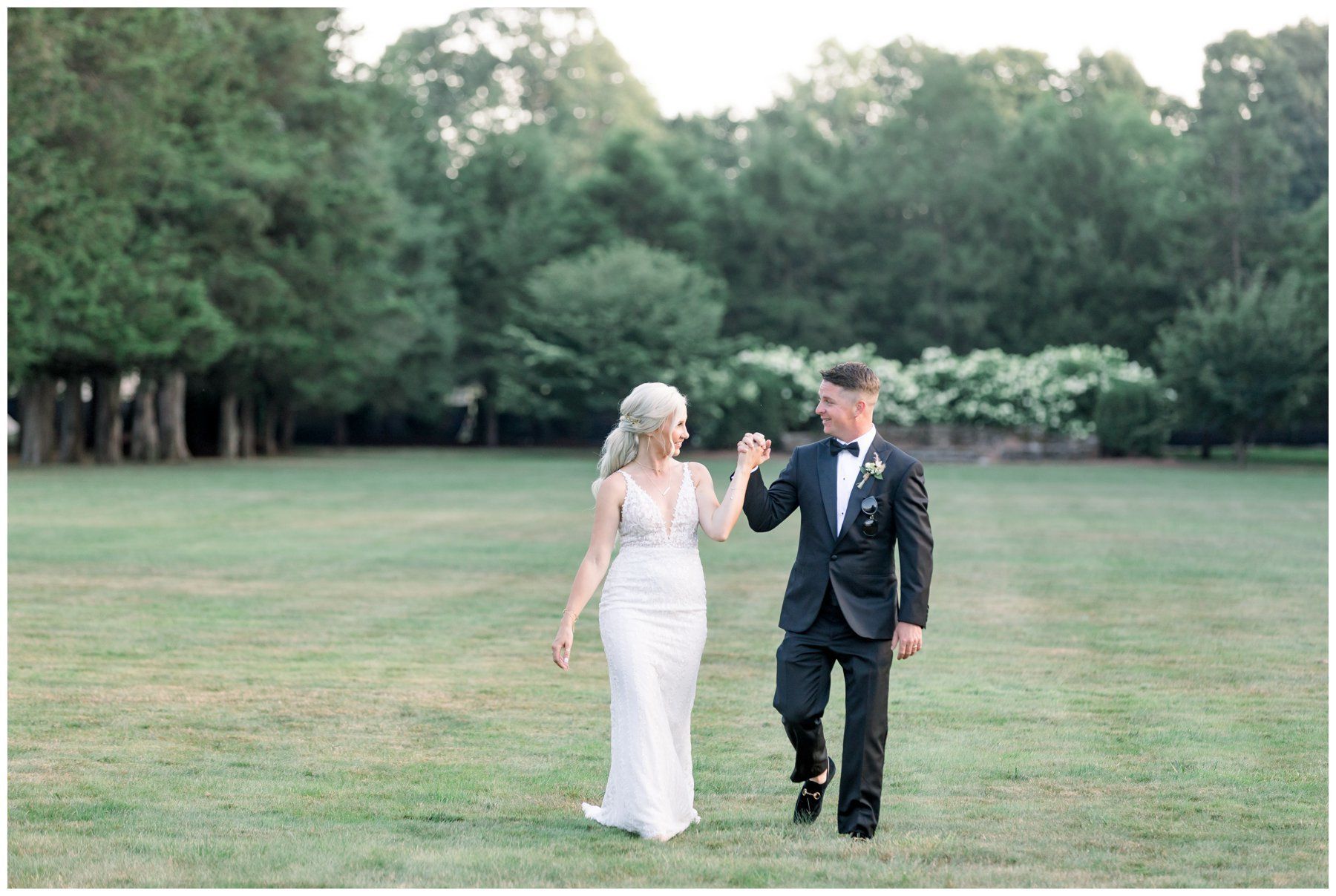 Two people in formal attire holding hands in a grassy field with trees in the background.