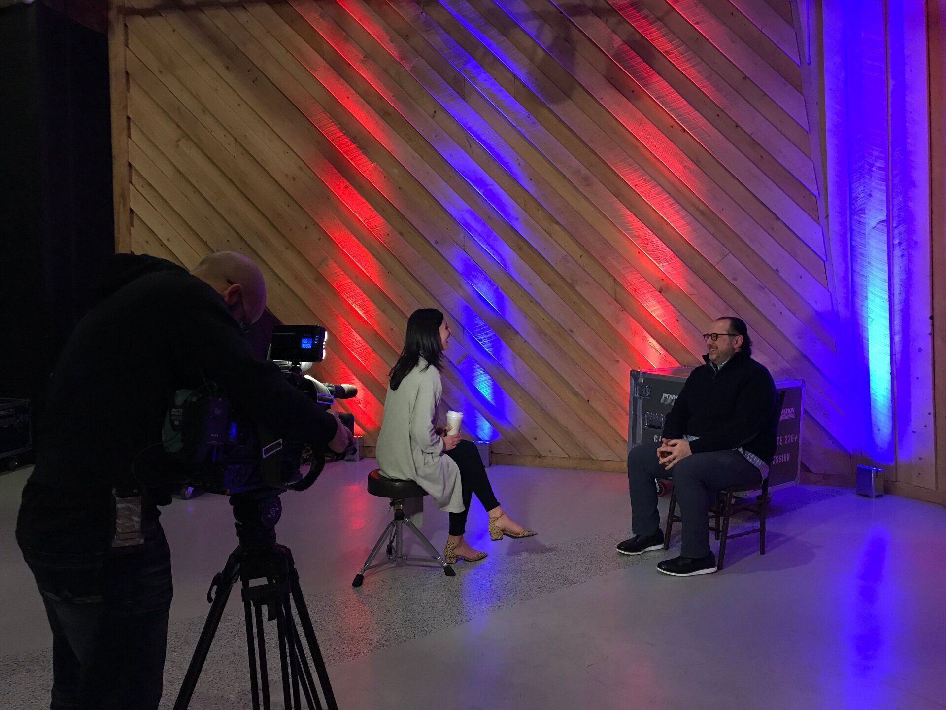 Studio interview set with two seated speakers, camera crew, and red and blue lighting backdrop
