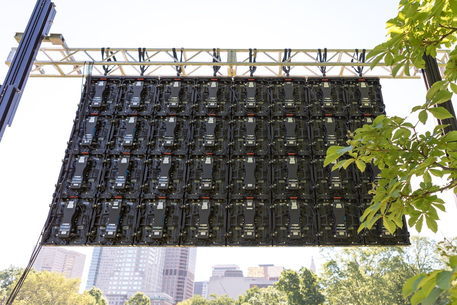 Large LED display wall suspended outdoors beneath a metal frame, with trees and city buildings in the background