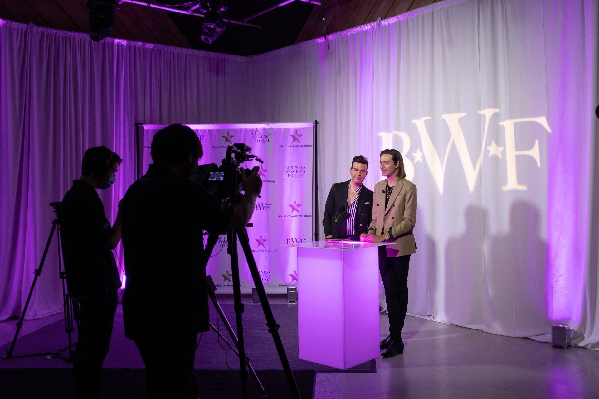 Two people at a lit podium in a purple-lit studio, with camera crew silhouettes filming nearby