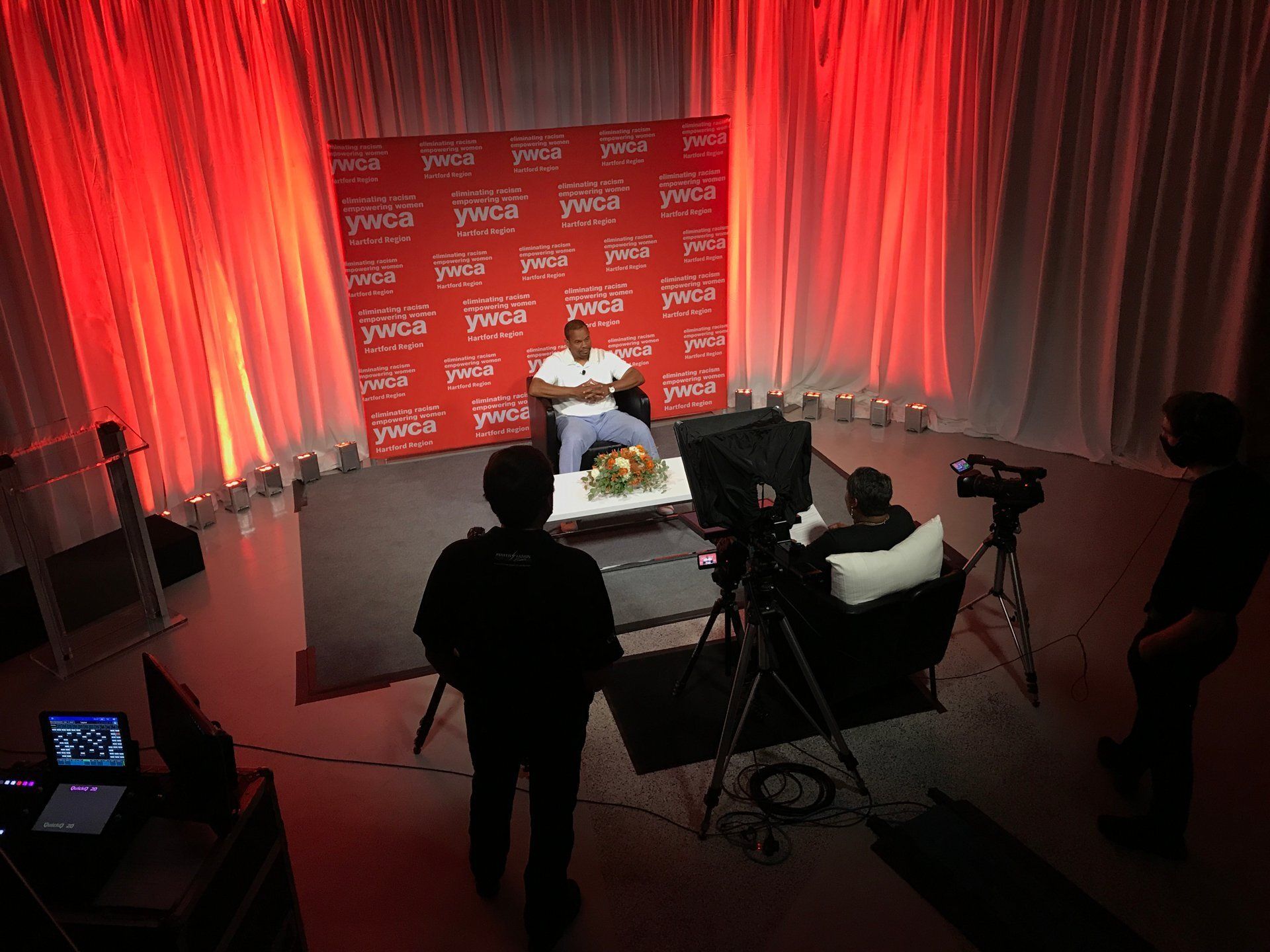 Interview set with a seated speaker, cameras, and red backdrop under studio lighting