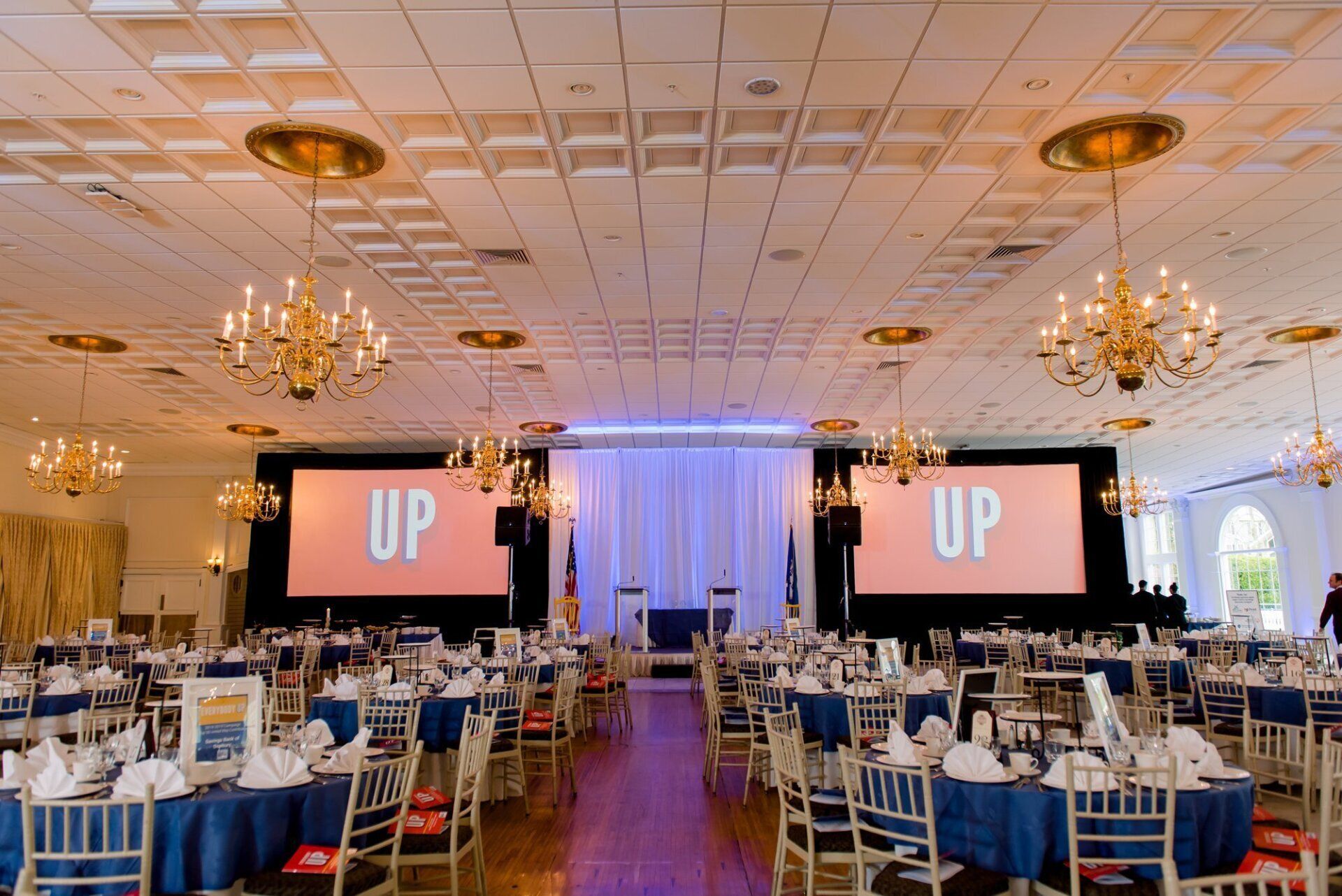 Banquet hall with blue table settings and two large screens showing “UP” at the front