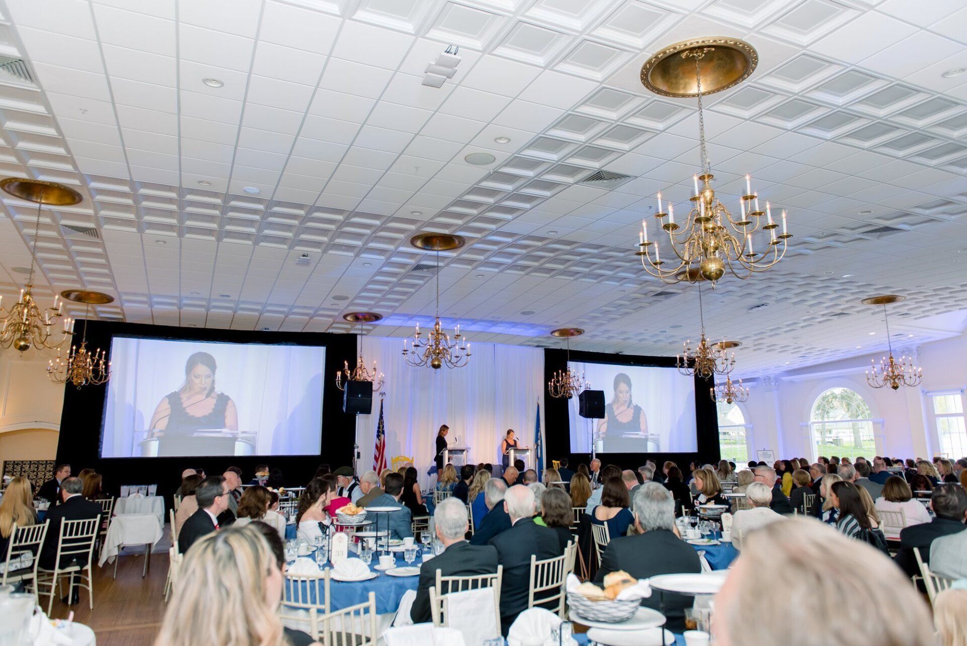 Banquet hall conference with seated audience and two large projection screens showing a speaker onstage