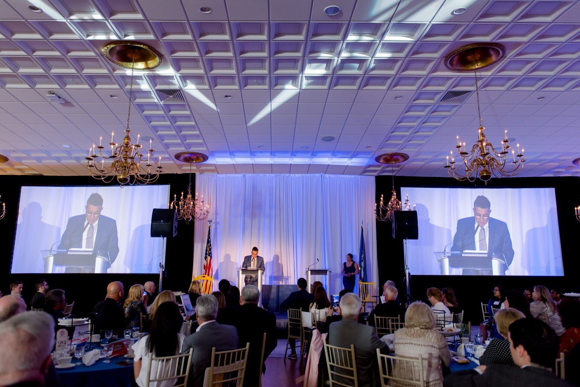 Conference banquet with a speaker on stage, projected on two large screens, audience seated at round tables under chandeliers