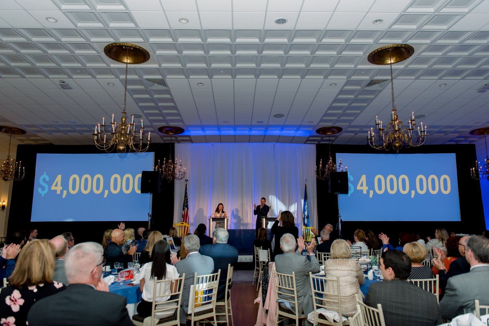 Audience at gala banquet watching speakers on stage with two screens showing “$4,000,000”