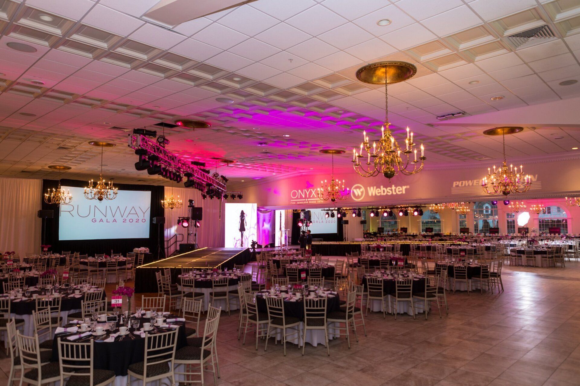 Banquet hall with round tables, chairs, pink lighting, and a stage screen at a wedding reception