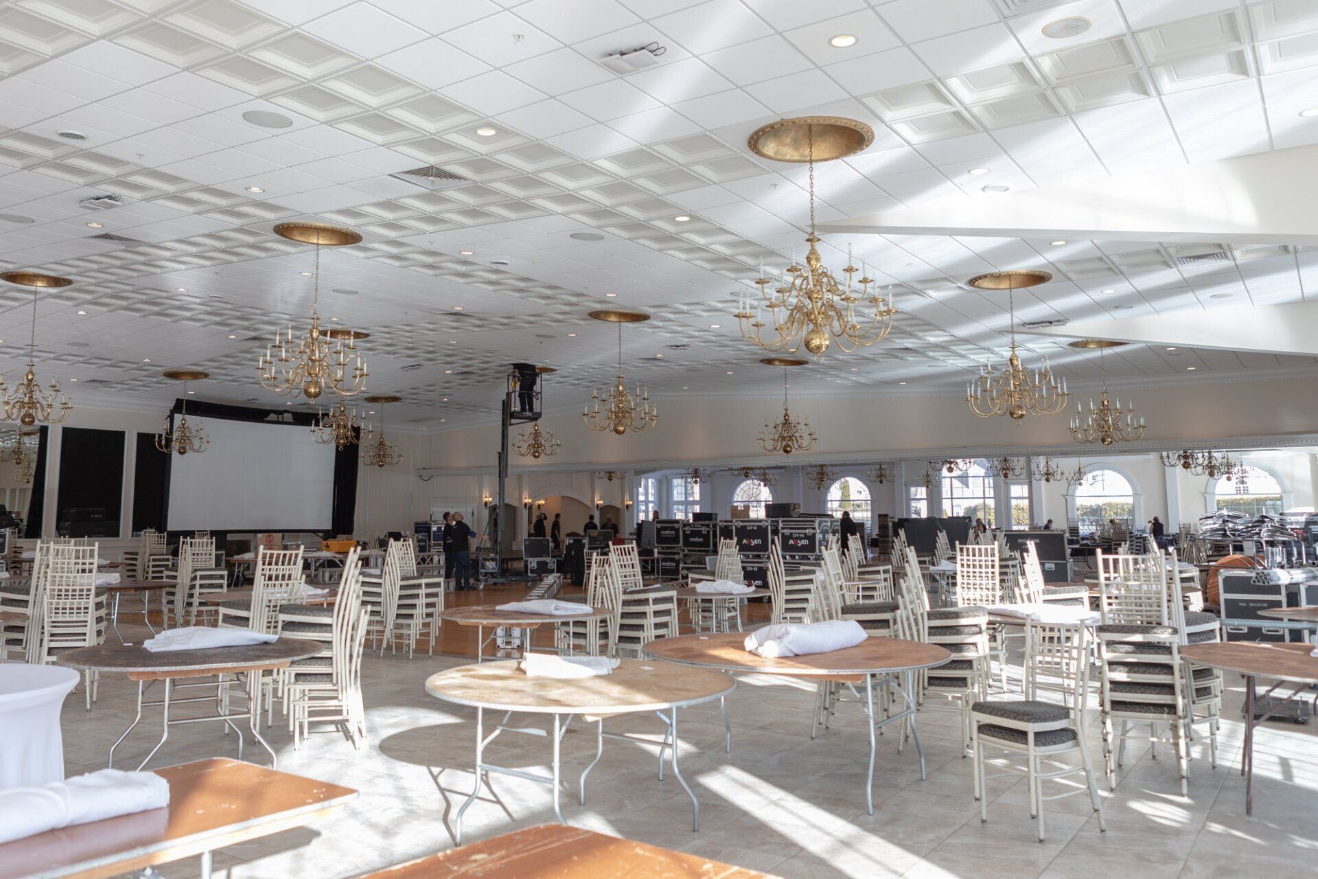 Empty banquet hall with white chairs, round tables, chandeliers, and a projection screen.
