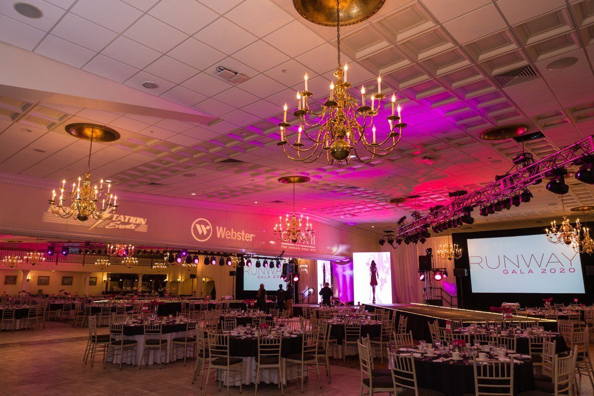 Banquet hall with pink lighting, chandeliers, tables, and a large runway stage for an event