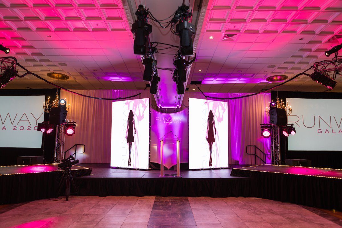 Elegant event runway with pink lighting, tall white screens, and purple drapes in a ballroom