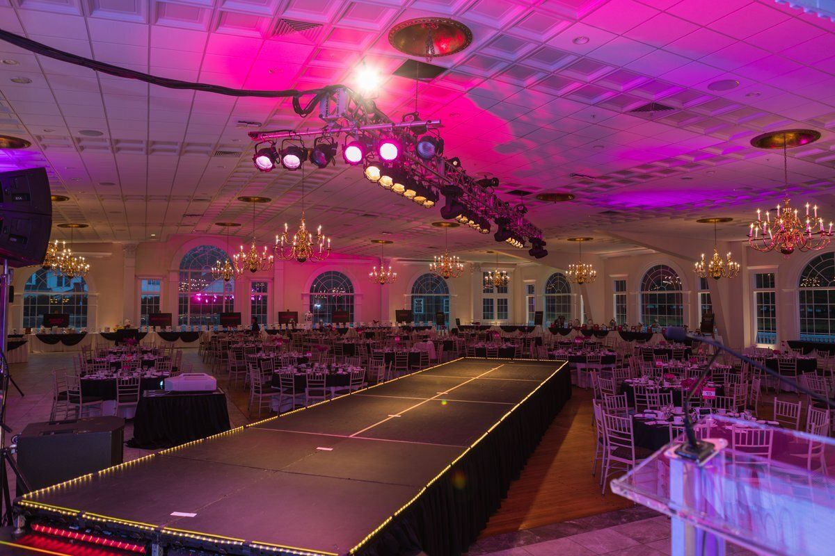 Elegant banquet hall with a lit runway, round tables, chandeliers, and pink-purple ceiling lighting