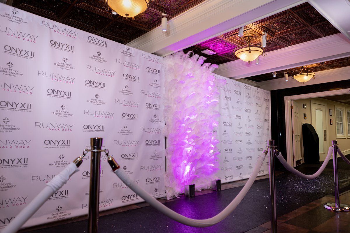 Roped-off event entrance with white step-and-repeat wall, purple uplighting, and ceiling chandeliers