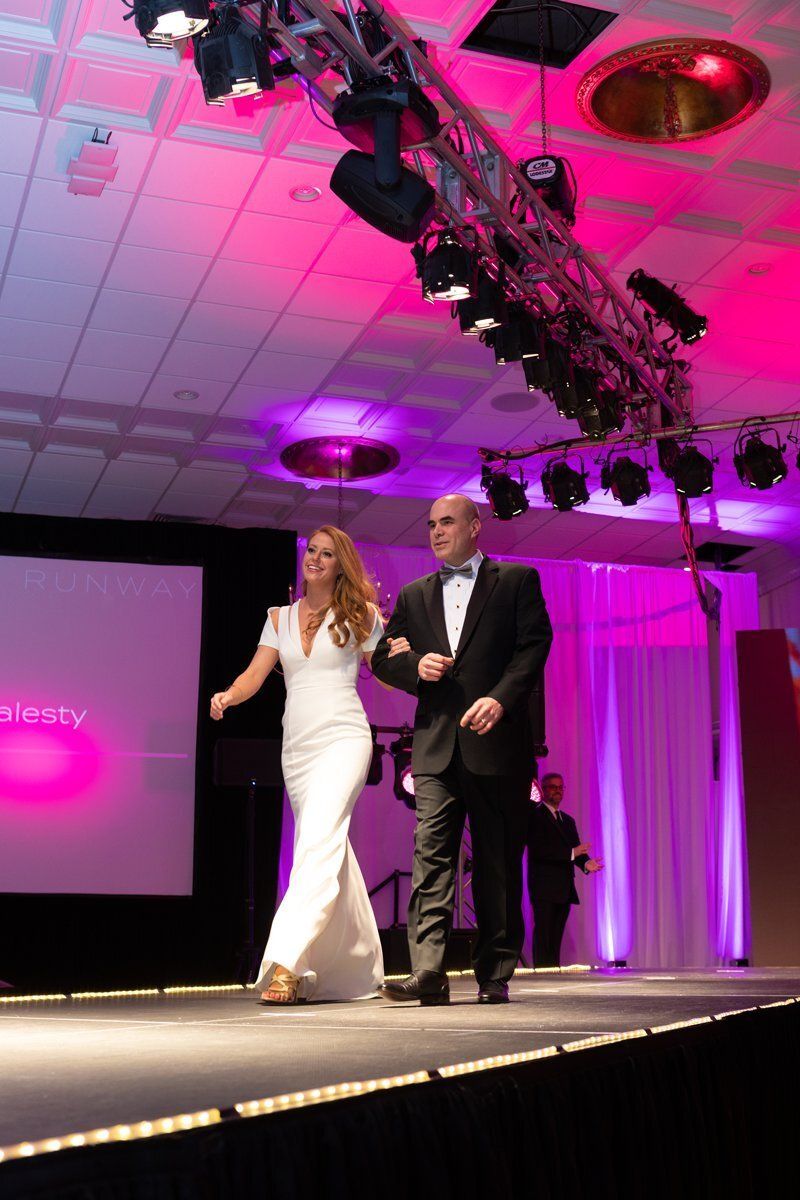 Two presenters walking on a runway stage under pink and purple lighting