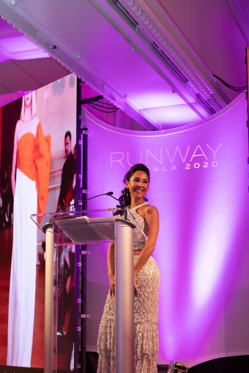 Woman in a sparkling gown speaking at a podium on a purple-lit runway stage