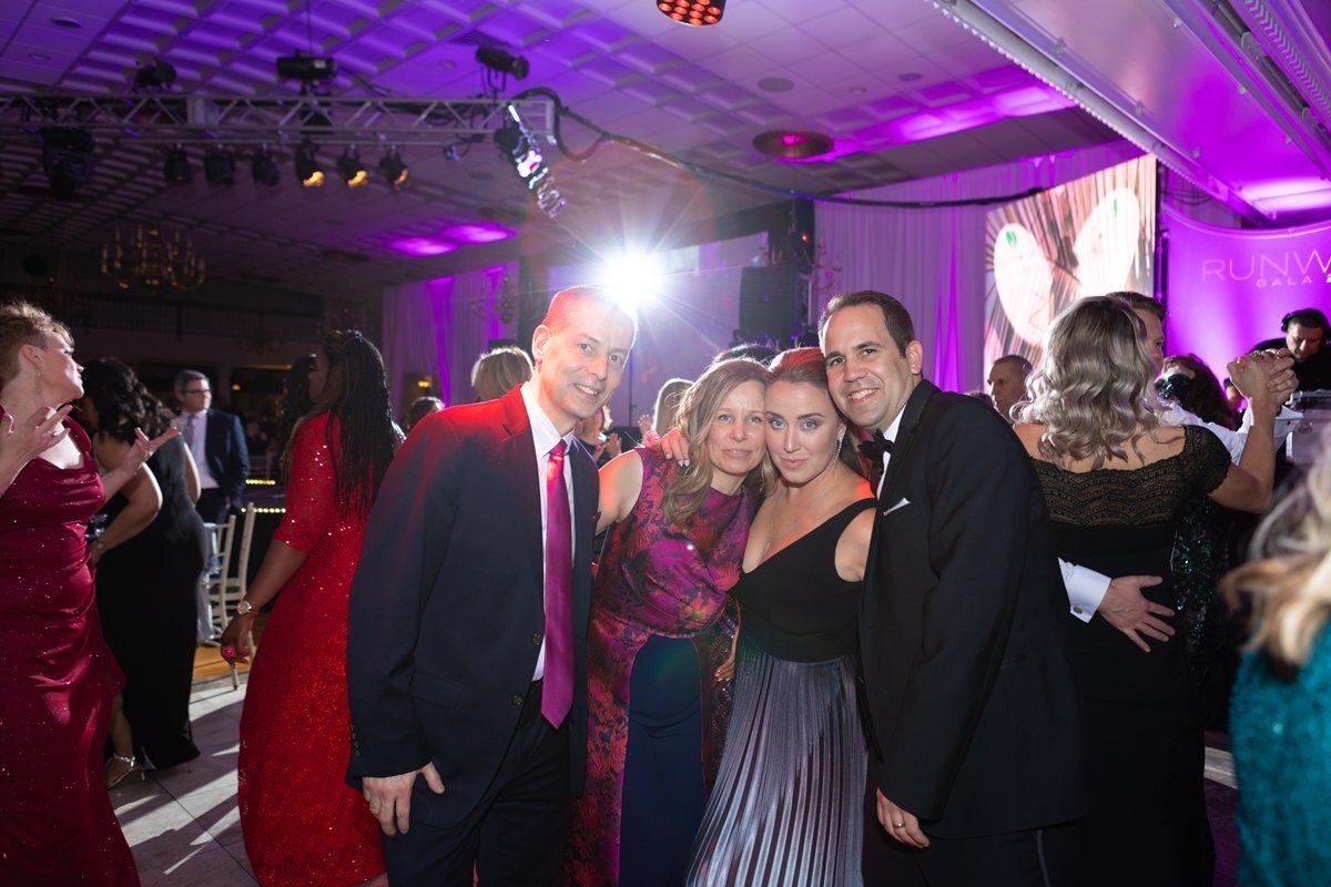 Guests posing at a formal party under purple lighting, with a bright camera flash in the background.