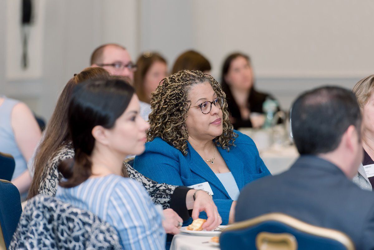 Audience listening at a conference table, with a woman in a blue blazer smiling in the foreground.