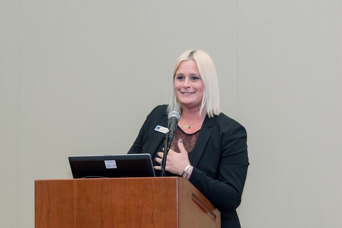Woman speaking at a podium with a microphone and laptop in a plain conference room.