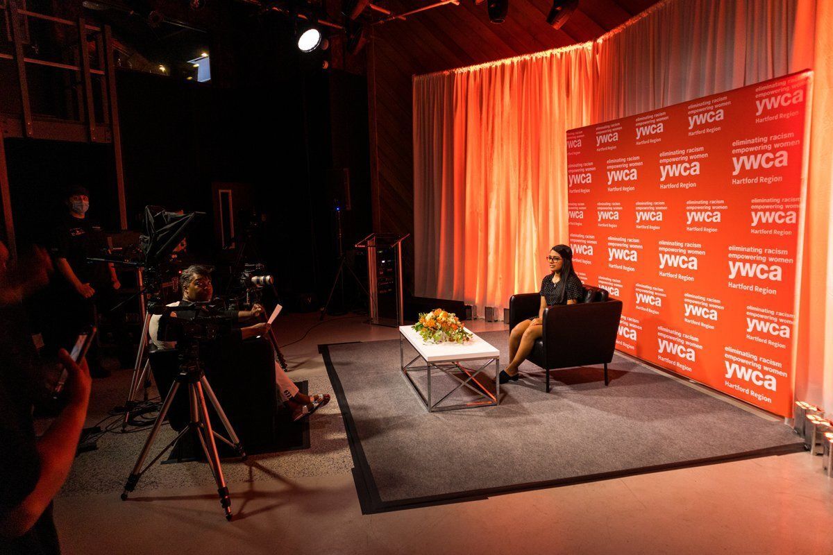 Interview set with seated person, cameras, lights, and orange sponsor backdrop on a rug.