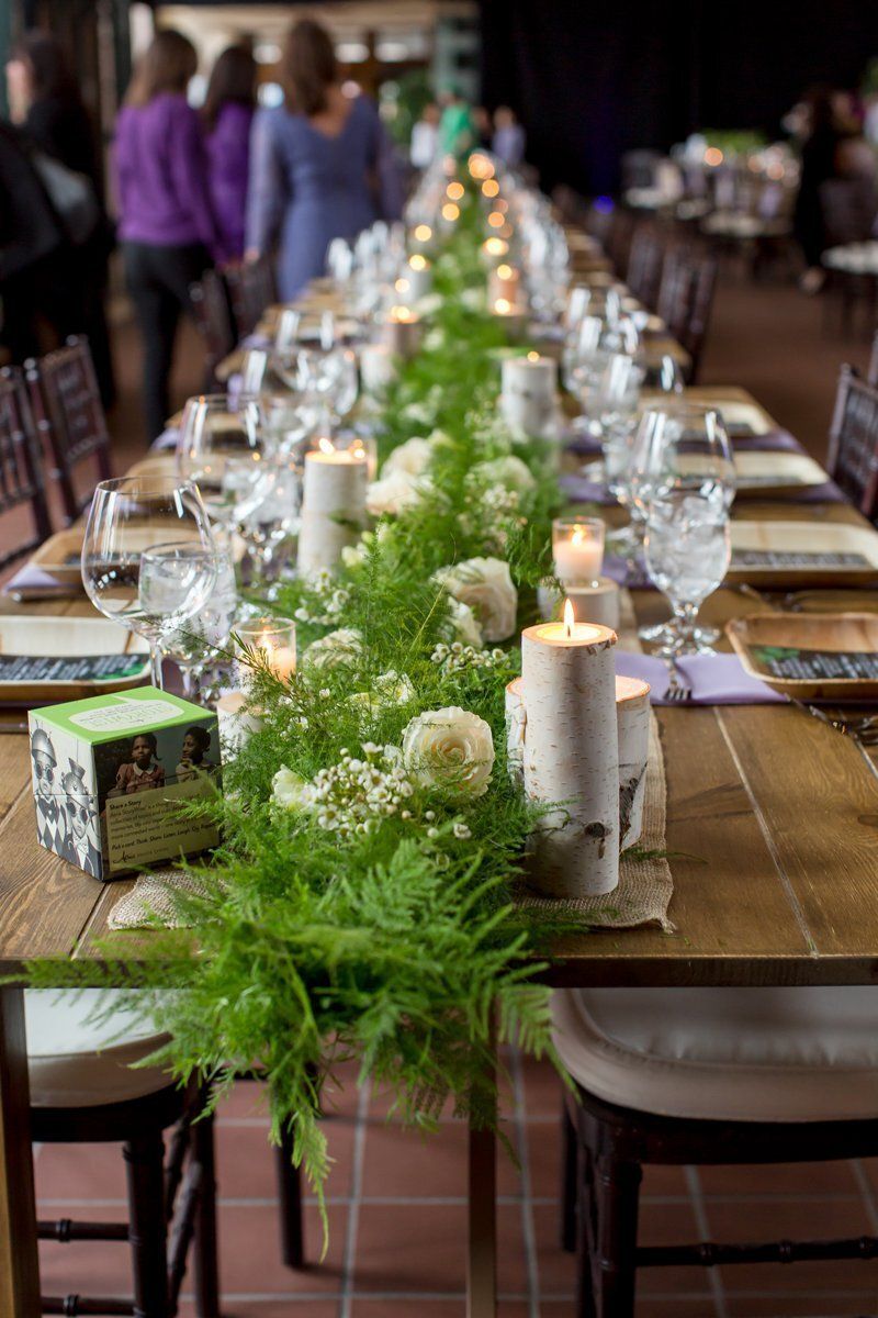 Long banquet table with glassware, candles, and greenery centerpieces in a warmly lit dining hall