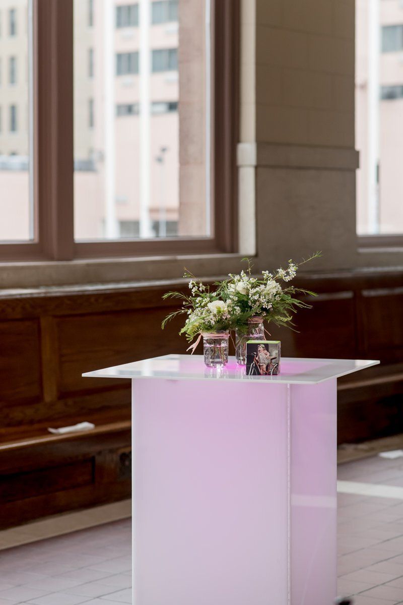 Small white table with pink lighting, two potted plants, and flowers in a bright room with windows.
