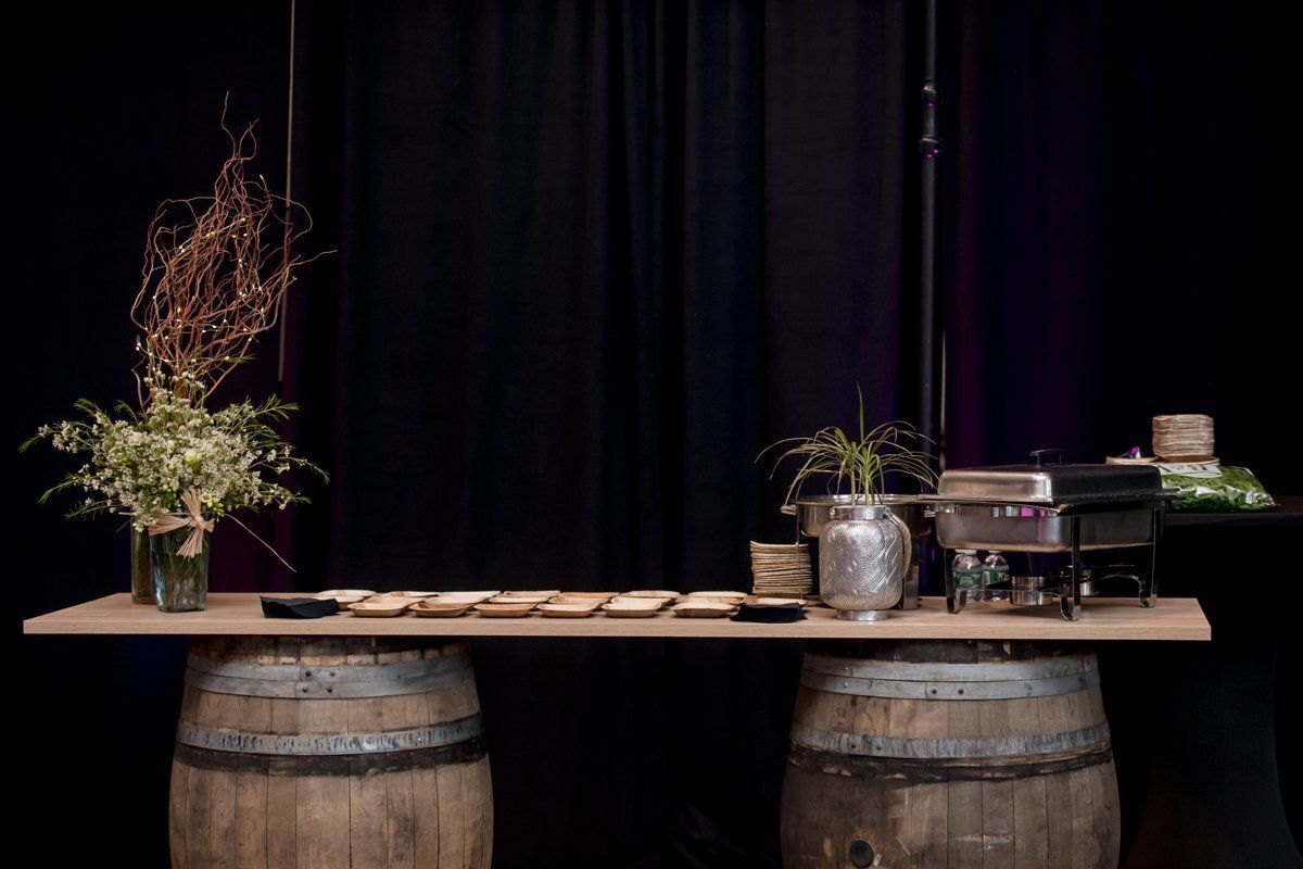 Rustic table setup with flowers, jars, and plates on barrel supports against a black curtain backdrop